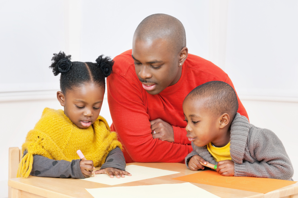 Two Black children sit at a table beside a Black man looking at worksheets. The children wear yellow attire and the man is in a red-orange shirt.