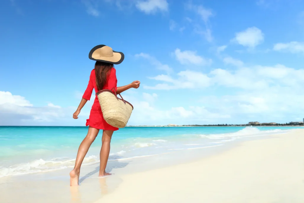 Female tourist in beach suit with straw sun hat and beach bag walking on tropical vacation, wearing sun hat and red tunic covering dress, relaxing on travel vacation from behind.  (Photo: Maridav / iStock / Getty Images Plus)