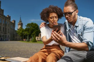 Young woman and man sitting on a bench and frowning while looking at the screen of a smartphone (Photo Credit: yacobchuk / iStock / Getty Images Plus)